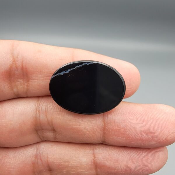 Multi-angle macro photographs of a 27.51-carat natural black banded agate (Sulemani Aqeeq) oval cabochon held in metal tweezers and fingertips, showcasing a mirror-polished deep obsidian black surface accented by fine, undulating white banding lines traversing across the polished surface.