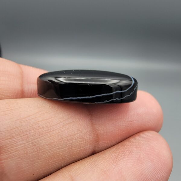 Multi-angle macro photographs of a 27.51-carat natural black banded agate (Sulemani Aqeeq) oval cabochon held in metal tweezers and fingertips, showcasing a mirror-polished deep obsidian black surface accented by fine, undulating white banding lines traversing across the polished surface.