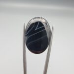 Multi-angle macro photographs of a 26.54-carat natural black banded agate (Sulemani Aqeeq) oval cabochon held vertically in metal tweezers, displaying a mirror-polished deep obsidian black surface accented by sharp V-shaped geometric chevron lines and fine parallel fortification banding.