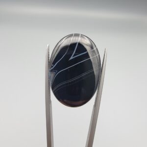 Multi-angle macro photographs of a 26.54-carat natural black banded agate (Sulemani Aqeeq) oval cabochon held vertically in metal tweezers, displaying a mirror-polished deep obsidian black surface accented by sharp V-shaped geometric chevron lines and fine parallel fortification banding.
