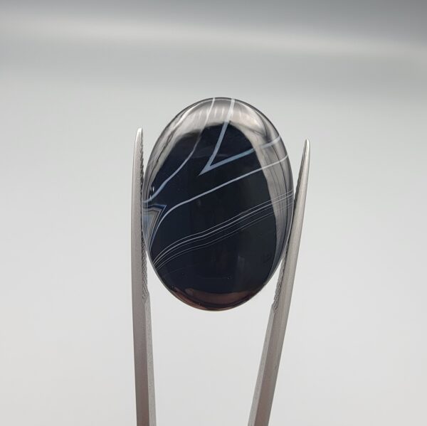 Multi-angle macro photographs of a 26.54-carat natural black banded agate (Sulemani Aqeeq) oval cabochon held vertically in metal tweezers, displaying a mirror-polished deep obsidian black surface accented by sharp V-shaped geometric chevron lines and fine parallel fortification banding.