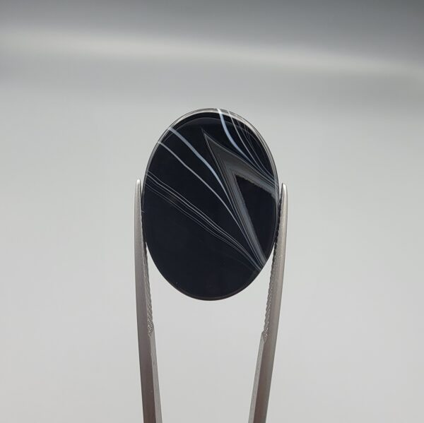Multi-angle macro photographs of a 26.54-carat natural black banded agate (Sulemani Aqeeq) oval cabochon held vertically in metal tweezers, displaying a mirror-polished deep obsidian black surface accented by sharp V-shaped geometric chevron lines and fine parallel fortification banding.