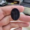 Multi-angle macro photographs of a 24.94-carat natural black banded agate (Sulemani Aqeeq) oval cabochon held in hand, displaying a mirror-polished deep obsidian black surface accented by sharp white angular geometric lines and fine parallel fortification banding across the face.