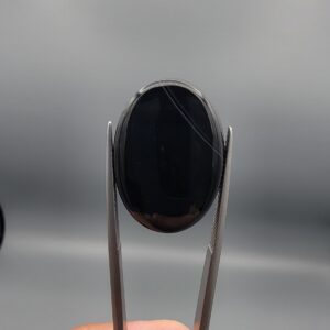 Multi-angle macro photographs of a 24.97-carat natural black banded agate (Sulemani Aqeeq) oval cabochon held in metal tweezers, displaying a mirror-polished deep obsidian black surface accented by crisp, fine diagonal white banding lines running across the face.
