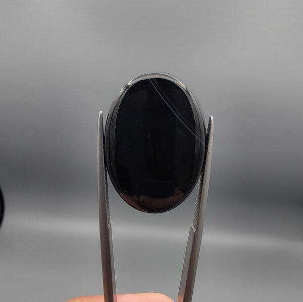 Multi-angle macro photographs of a 24.97-carat natural black banded agate (Sulemani Aqeeq) oval cabochon held in metal tweezers, displaying a mirror-polished deep obsidian black surface accented by crisp, fine diagonal white banding lines running across the face.
