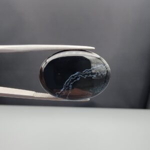 Multi-angle macro photographs of a 27.16-carat natural black banded agate (Sulemani Aqeeq) oval cabochon held in metal tweezers, showcasing an intense mirror-polished deep obsidian black surface accented by a meandering white scalloped or lace-like banding arc across the face.