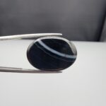 Multi-angle macro photographs of a 26.14-carat natural black banded agate (Sulemani Aqeeq) oval cabochon held in metal tweezers, showcasing a mirror-polished deep obsidian black dome with prominent, gently curved white banding lines traversing across the polished surface.