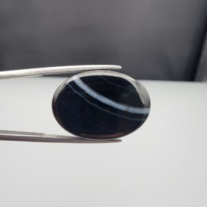 Multi-angle macro photographs of a 26.14-carat natural black banded agate (Sulemani Aqeeq) oval cabochon held in metal tweezers, showcasing a mirror-polished deep obsidian black dome with prominent, gently curved white banding lines traversing across the polished surface.