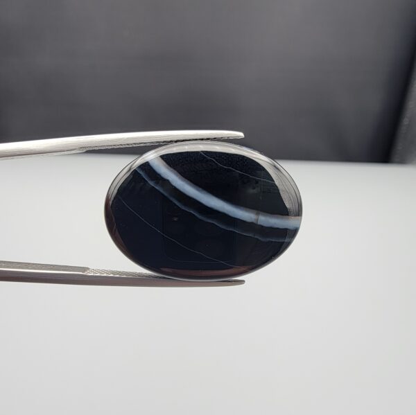Multi-angle macro photographs of a 26.14-carat natural black banded agate (Sulemani Aqeeq) oval cabochon held in metal tweezers, showcasing a mirror-polished deep obsidian black dome with prominent, gently curved white banding lines traversing across the polished surface.