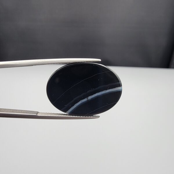 Multi-angle macro photographs of a 26.14-carat natural black banded agate (Sulemani Aqeeq) oval cabochon held in metal tweezers, showcasing a mirror-polished deep obsidian black dome with prominent, gently curved white banding lines traversing across the polished surface.