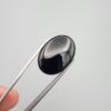 Multi-angle macro photographs of a 26.84-carat natural black banded agate (Sulemani Aqeeq) oval cabochon held in metal tweezers, showcasing a mirror-polished deep obsidian black surface accented by a single, distinct curved white banding line along the right edge.