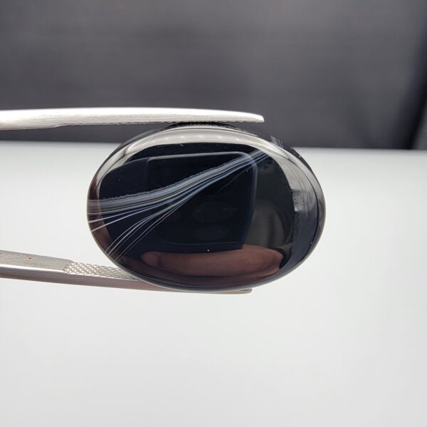 Multi-angle macro photographs of a 25.17-carat natural black banded agate (Sulemani Aqeeq) oval cabochon held in metal tweezers, showcasing a mirror-polished deep obsidian black surface accented by multiple fine, sharp parallel white banding lines sweeping diagonally across the face.
