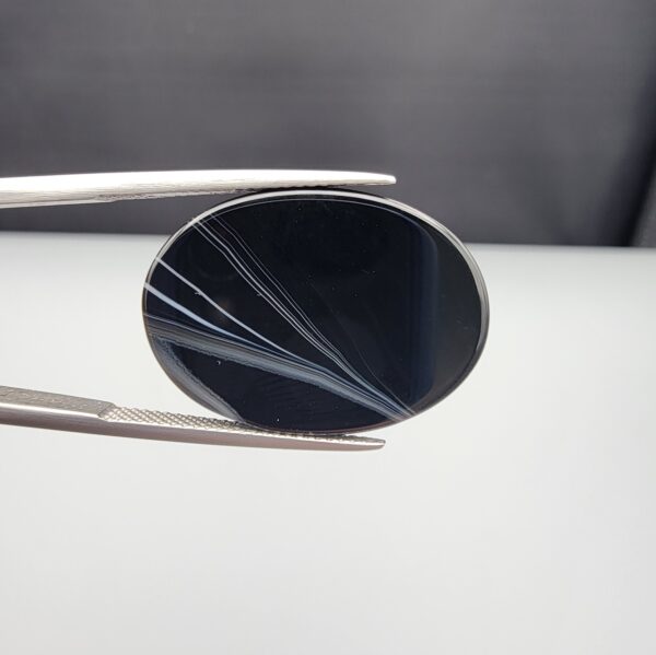 Multi-angle macro photographs of a 25.17-carat natural black banded agate (Sulemani Aqeeq) oval cabochon held in metal tweezers, showcasing a mirror-polished deep obsidian black surface accented by multiple fine, sharp parallel white banding lines sweeping diagonally across the face.
