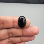 Multi-angle macro photographs of a 10.41-carat natural black banded agate (Sulemani Aqeeq) oval cabochon held on fingertips, showcasing a mirror-polished obsidian black surface contrasting with a wide, sweeping curved band of luminous blue-grey and white forming a bold eye-like pattern.