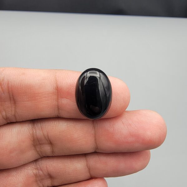 Multi-angle macro photographs of a 10.41-carat natural black banded agate (Sulemani Aqeeq) oval cabochon held on fingertips, showcasing a mirror-polished obsidian black surface contrasting with a wide, sweeping curved band of luminous blue-grey and white forming a bold eye-like pattern.