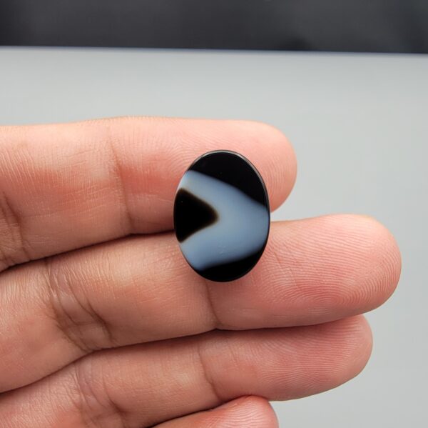 Multi-angle macro photographs of a 10.41-carat natural black banded agate (Sulemani Aqeeq) oval cabochon held on fingertips, showcasing a mirror-polished obsidian black surface contrasting with a wide, sweeping curved band of luminous blue-grey and white forming a bold eye-like pattern.