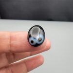 Multi-angle macro photographs of a 9.91-carat natural black banded agate (Sulemani Aqeeq) oval cabochon held on fingertips, displaying a mirror-polished deep obsidian black dome featuring a distinct dual "eye" pattern composed of concentric blue-grey and white rings around dark centers at one end.