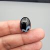 Multi-angle macro photographs of a 9.91-carat natural black banded agate (Sulemani Aqeeq) oval cabochon held on fingertips, displaying a mirror-polished deep obsidian black dome featuring a distinct dual "eye" pattern composed of concentric blue-grey and white rings around dark centers at one end.