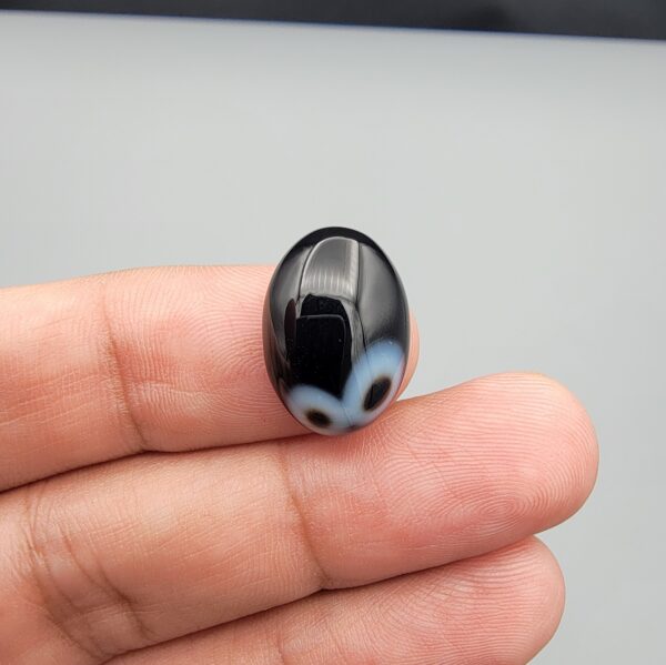 Multi-angle macro photographs of a 9.91-carat natural black banded agate (Sulemani Aqeeq) oval cabochon held on fingertips, displaying a mirror-polished deep obsidian black dome featuring a distinct dual "eye" pattern composed of concentric blue-grey and white rings around dark centers at one end.