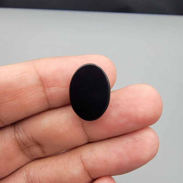 Multi-angle macro photographs of a 9.91-carat natural black banded agate (Sulemani Aqeeq) oval cabochon held on fingertips, displaying a mirror-polished deep obsidian black dome featuring a distinct dual "eye" pattern composed of concentric blue-grey and white rings around dark centers at one end.