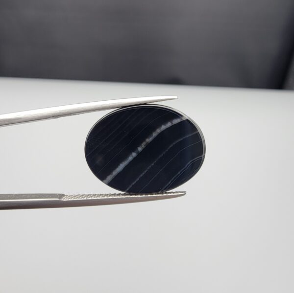 Multi-angle macro photographs of a 15.74-carat natural black banded agate (Sulemani Aqeeq) oval cabochon held in metal tweezers, showcasing a mirror-polished deep obsidian black surface accented by clean, parallel diagonal white banding lines running across the face.