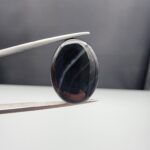 Multi-angle macro photographs of a 15.74-carat natural black banded agate (Sulemani Aqeeq) oval cabochon held in metal tweezers, showcasing a mirror-polished deep obsidian black surface accented by clean, parallel diagonal white banding lines running across the face.