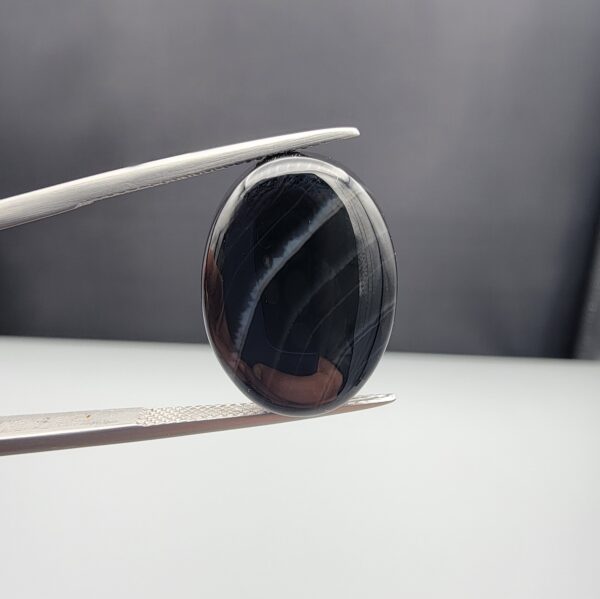 Multi-angle macro photographs of a 15.74-carat natural black banded agate (Sulemani Aqeeq) oval cabochon held in metal tweezers, showcasing a mirror-polished deep obsidian black surface accented by clean, parallel diagonal white banding lines running across the face.