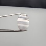 Multi-angle macro photographs of an 18.37-carat natural banded agate (Sulemani Aqeeq) oval cabochon held in metal tweezers, showcasing a smooth, high-gloss surface, translucent monochrome grey-and-white horizontal banding, and fine natural mineral strata.