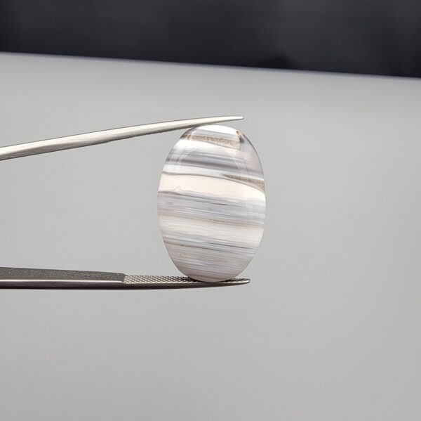 Multi-angle macro photographs of an 18.37-carat natural banded agate (Sulemani Aqeeq) oval cabochon held in metal tweezers, showcasing a smooth, high-gloss surface, translucent monochrome grey-and-white horizontal banding, and fine natural mineral strata.