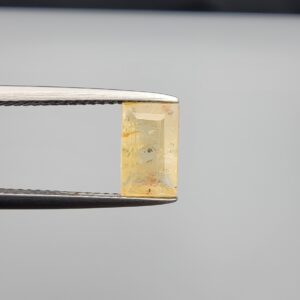 Macro photograph of a 2.12-carat natural yellow sapphire (Pukhraj) rectangular step-cut gemstone from Sri Lanka held in metal tweezers, showcasing crisp geometry, a warm light-golden yellow hue, and authentic unheated internal mineral inclusions.