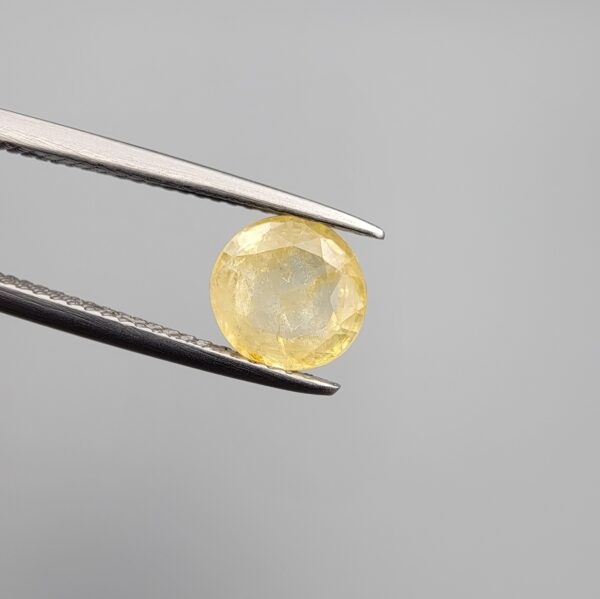 Macro photograph of a 2.12-carat natural pale yellow sapphire (Pukhraj) round mixed-cut gemstone from Sri Lanka held in metal tweezers, showcasing a delicate straw-yellow hue, precision faceting, and natural unheated crystalline internal texture.