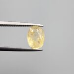Macro photograph of a 2.85-carat natural pale yellow sapphire (Pukhraj) oval mixed-cut gemstone from Sri Lanka held in metal tweezers, showcasing a delicate lemon-gold hue, precision faceting, and authentic unheated internal crystalline texture.