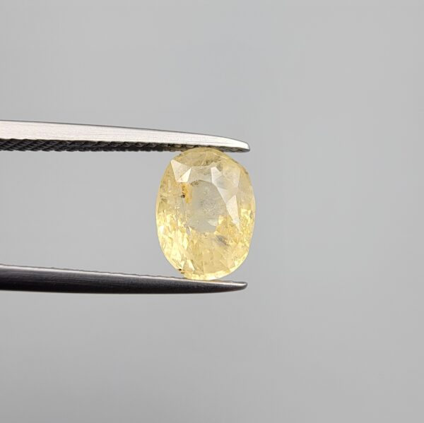Macro photograph of a 2.85-carat natural pale yellow sapphire (Pukhraj) oval mixed-cut gemstone from Sri Lanka held in metal tweezers, showcasing a delicate lemon-gold hue, precision faceting, and authentic unheated internal crystalline texture.