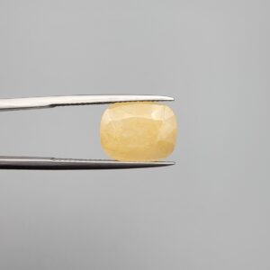 Macro photograph of an 8.45-carat natural yellow sapphire (Pukhraj) cushion-cut gemstone from Sri Lanka held in metal tweezers, showcasing precision faceting, a warm lemon-golden hue, and authentic unheated clarity.