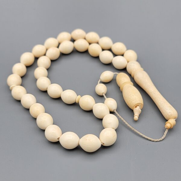 High-resolution macro photograph of a 63.47-gram handmade natural camel bone 33-bead Islamic Tasbih (prayer beads) laid out on a neutral studio backdrop, showcasing smooth ivory-hued barley-cut bone beads, carved separators, and a masterfully turned main pillar (imame).