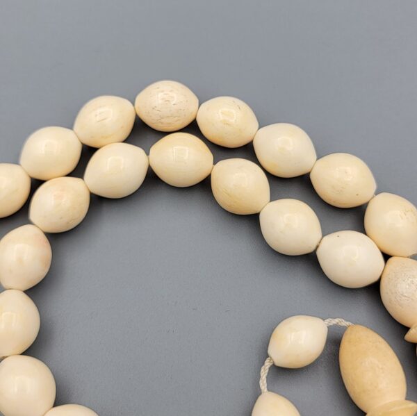 High-resolution macro photograph of a 63.47-gram handmade natural camel bone 33-bead Islamic Tasbih (prayer beads) laid out on a neutral studio backdrop, showcasing smooth ivory-hued barley-cut bone beads, carved separators, and a masterfully turned main pillar (imame).