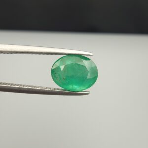 Macro photograph of a 2.19-carat natural Zambian emerald (Zamrud) held in metal tweezers, displaying a faceted oval cut, vivid green saturation, and fine natural internal inclusions (jardin) across the crown.