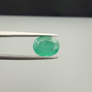 Macro photograph of a 1.91-carat natural Zambian emerald (Zamrud) held in metal tweezers, showcasing a faceted oval cut, vivid green hue, and natural internal crystal growth inclusions (jardin) across the crown.