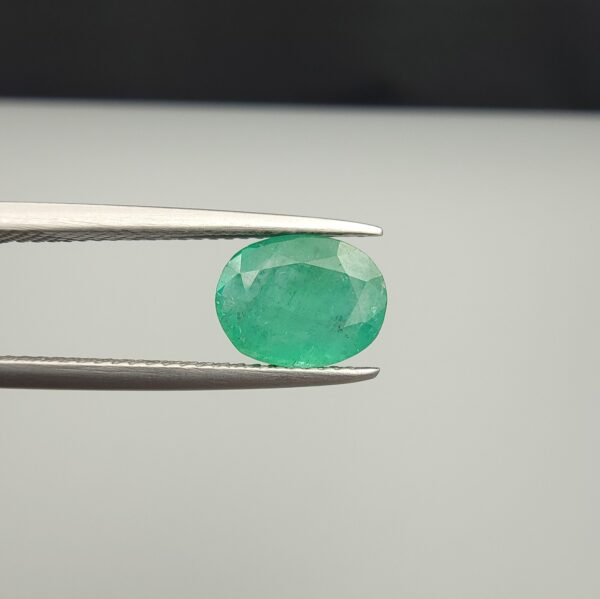 Macro photograph of a 1.91-carat natural Zambian emerald (Zamrud) held in metal tweezers, showcasing a faceted oval cut, vivid green hue, and natural internal crystal growth inclusions (jardin) across the crown.