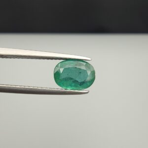 Macro photograph of a 1.71-carat natural Zambian emerald (Zamrud) held horizontally in stainless steel gem tweezers, displaying a faceted oval cut, deep rich green color, and fine natural internal inclusions (jardin) across the table.