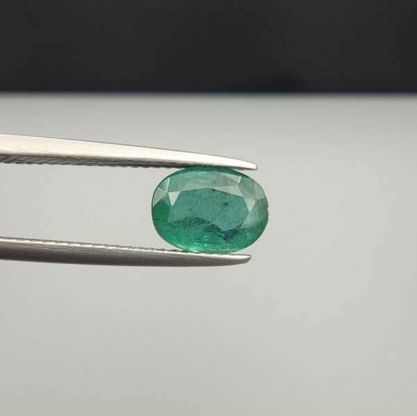 Macro photograph of a 1.71-carat natural Zambian emerald (Zamrud) held horizontally in stainless steel gem tweezers, displaying a faceted oval cut, deep rich green color, and fine natural internal inclusions (jardin) across the table.