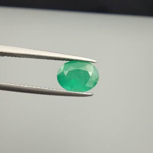 Macro photograph of a 1.79-carat natural Zambian emerald (Zamrud) held in metal tweezers, showcasing a faceted oval cut, vivid green saturation, and fine natural internal inclusions (jardin) across the table.