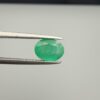 Macro photograph of a 1.51-carat natural Zambian emerald (Zamrud) held in metal gem tweezers, displaying a faceted oval cut, vivid green saturation, and fine natural internal inclusions (jardin) across the table.