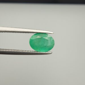 Macro photograph of a 1.51-carat natural Zambian emerald (Zamrud) held in metal gem tweezers, displaying a faceted oval cut, vivid green saturation, and fine natural internal inclusions (jardin) across the table.