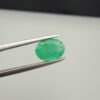 Macro photograph of a 1.51-carat natural Zambian emerald (Zamrud) held in metal gem tweezers, displaying a faceted oval cut, vivid green saturation, and fine natural internal inclusions (jardin) across the table.