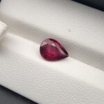 High-resolution macro photograph of a 2.02-carat natural Madagascar Rhodolite Garnet gemstone set against a white textured leather backdrop, displaying a brilliantly faceted pear cut with rich, saturated raspberry pinkish-red hues and exceptional transparency.