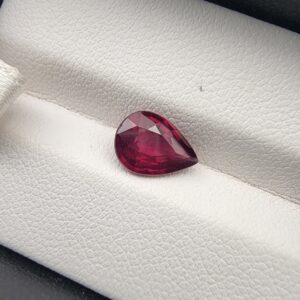 High-resolution macro photograph of a 2.02-carat natural Madagascar Rhodolite Garnet gemstone set against a white textured leather backdrop, displaying a brilliantly faceted pear cut with rich, saturated raspberry pinkish-red hues and exceptional transparency.