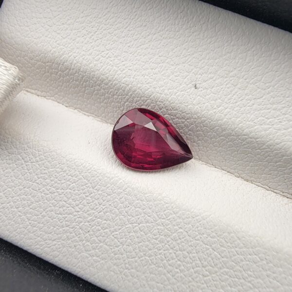 High-resolution macro photograph of a 2.02-carat natural Madagascar Rhodolite Garnet gemstone set against a white textured leather backdrop, displaying a brilliantly faceted pear cut with rich, saturated raspberry pinkish-red hues and exceptional transparency.