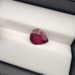 High-resolution macro photograph of a 3.17-carat natural Madagascar Rhodolite Garnet gemstone set against a white textured leather display cushion, displaying a brilliantly faceted pear cut with rich, saturated raspberry-red coloration and deep purplish-pink flashes.