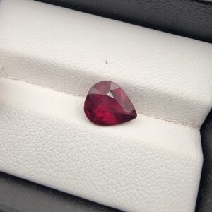 High-resolution macro photograph of a 3.17-carat natural Madagascar Rhodolite Garnet gemstone set against a white textured leather display cushion, displaying a brilliantly faceted pear cut with rich, saturated raspberry-red coloration and deep purplish-pink flashes.